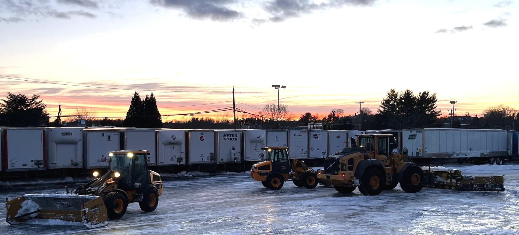 Three Coastal Corp wheel loaders with plows staged in a trailer yard at dawn after an overnight storm.