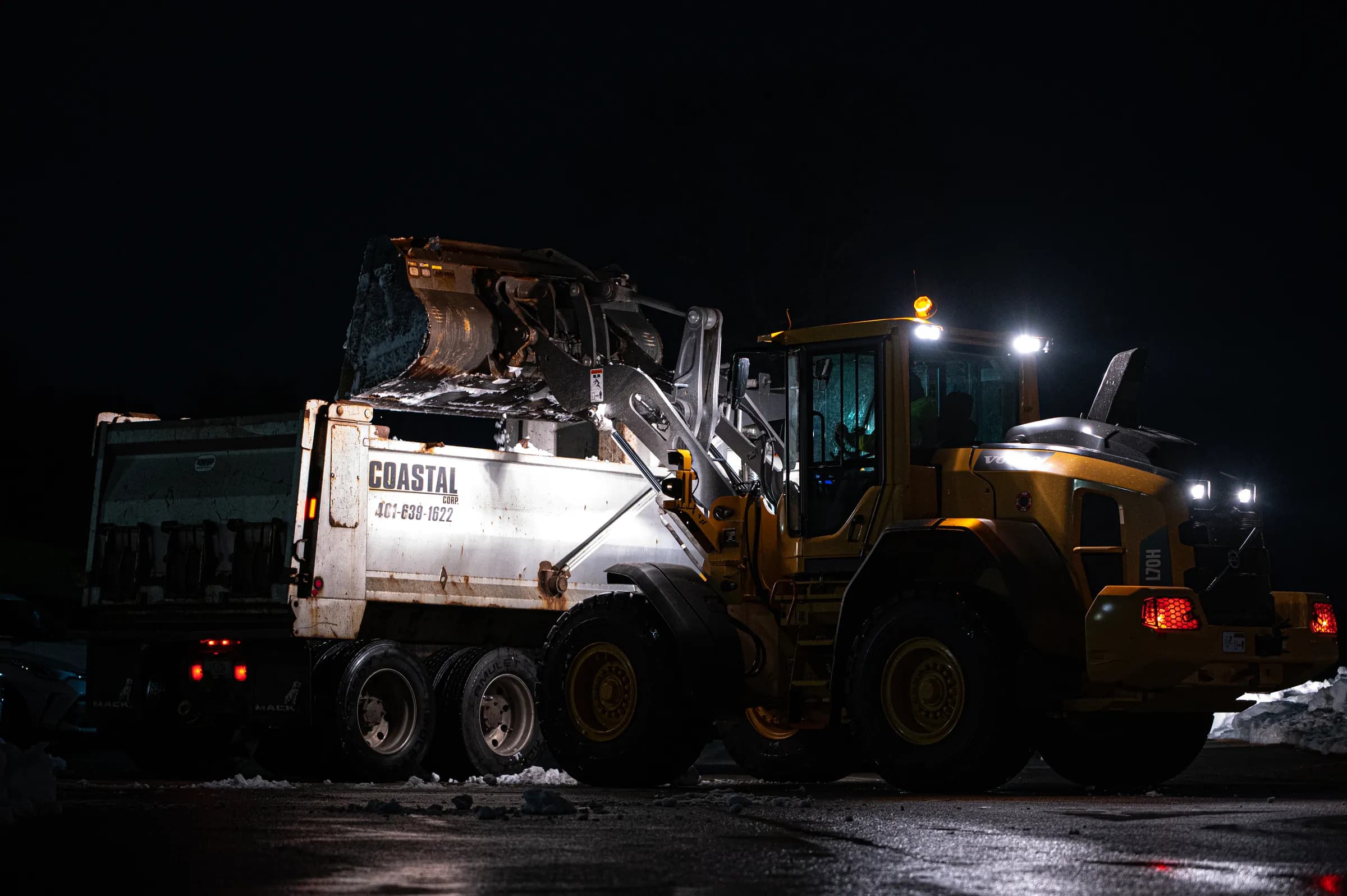 A Coastal Corp wheel loader loads snow into a Coastal dump truck during an overnight commercial clearing operation.