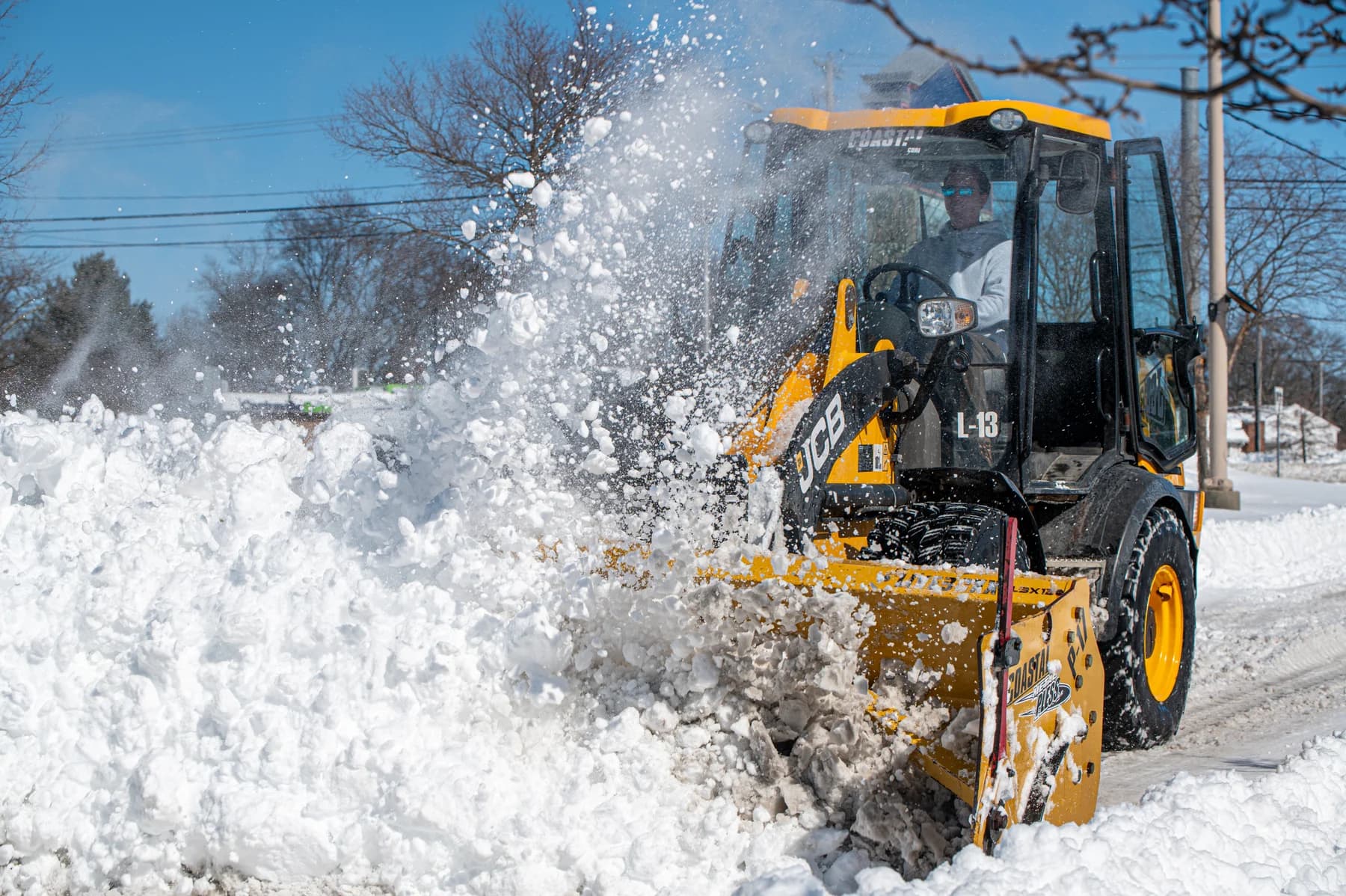 A Coastal Corp JCB loader throws a wide arc of snow while clearing a commercial roadway in daylight.