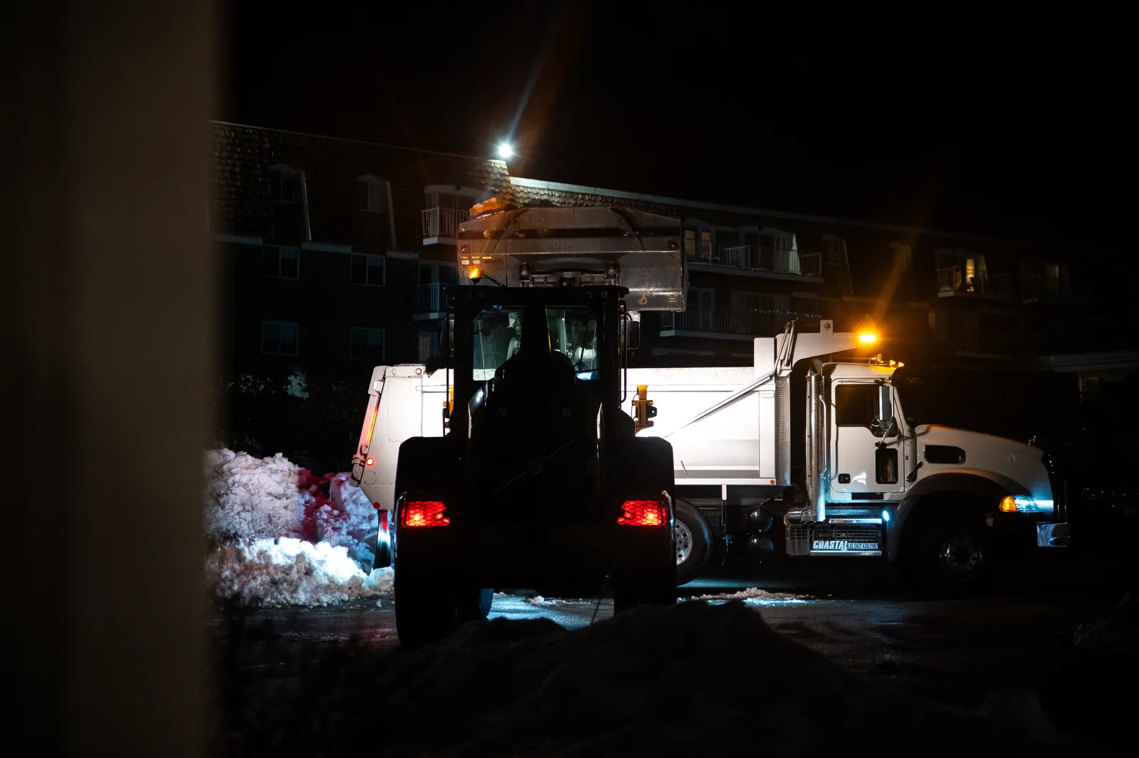 A Coastal Corp wheel loader and Mack dump truck clear and haul snow from a commercial complex at night.